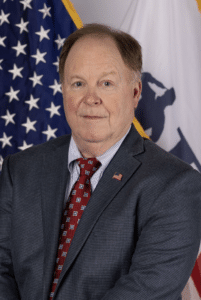 A man with light skin, short hair, and a receding hairline is wearing a gray patterned suit, a red patterned tie, and an American flag pin. He is standing in front of the U.S. flag and another flag with a partial emblem visible.